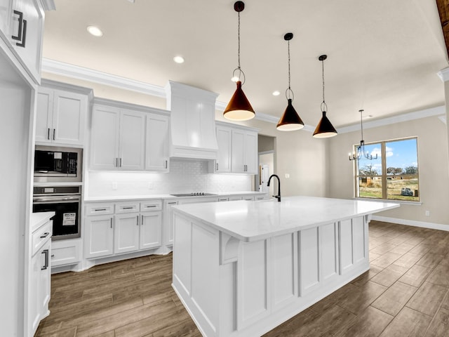 kitchen featuring stainless steel oven, decorative light fixtures, an island with sink, built in microwave, and white cabinets