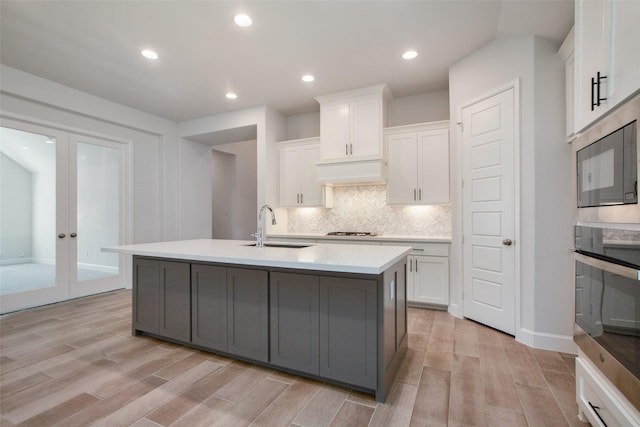 kitchen with stainless steel oven, french doors, a kitchen island with sink, white cabinetry, and sink