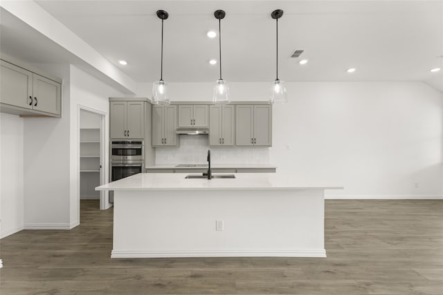 kitchen featuring gray cabinetry, hanging light fixtures, an island with sink, and stainless steel double oven