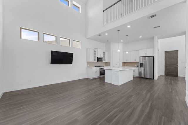 kitchen featuring stainless steel appliances, decorative light fixtures, white cabinets, a towering ceiling, and an island with sink