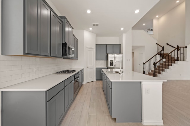 kitchen with gray cabinetry, stainless steel appliances, light hardwood / wood-style floors, sink, and a center island with sink