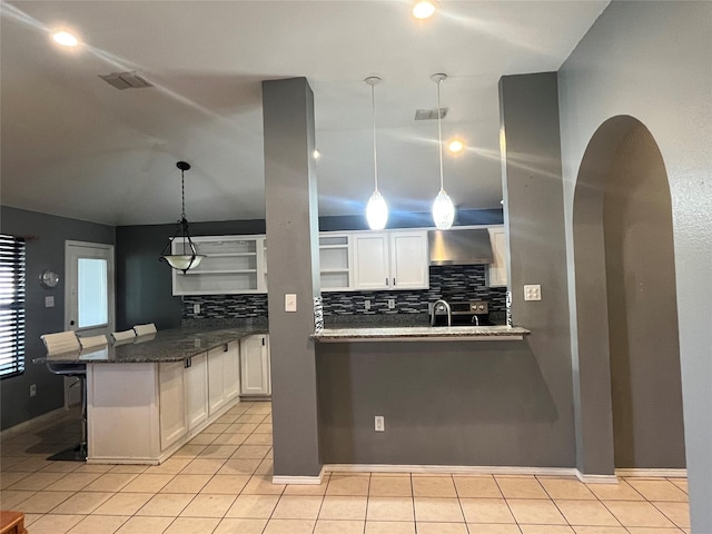 kitchen featuring decorative light fixtures, wall chimney range hood, white cabinetry, dark stone countertops, and a kitchen breakfast bar