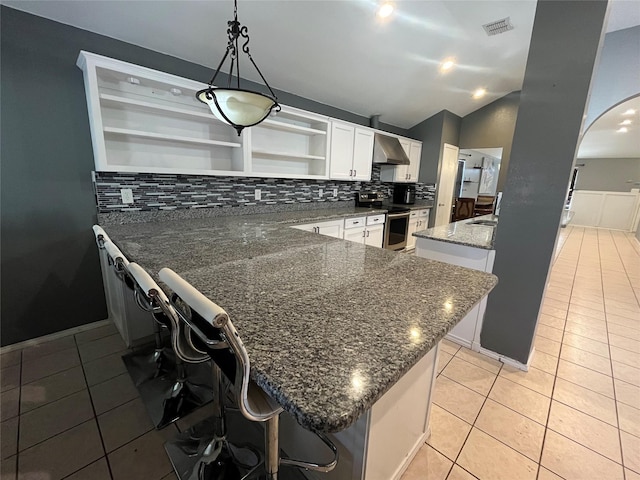 kitchen with stainless steel range with electric cooktop, white cabinetry, a kitchen bar, dark stone counters, and wall chimney range hood