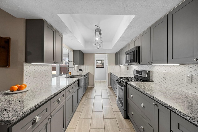 kitchen with a raised ceiling, light stone counters, appliances with stainless steel finishes, gray cabinetry, and a sink