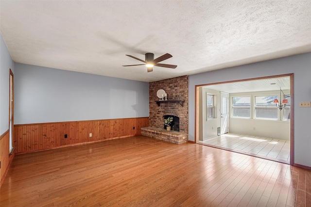 unfurnished living room with a textured ceiling, ceiling fan, light wood-type flooring, wainscoting, and a brick fireplace