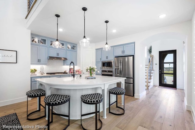 kitchen with stainless steel appliances, a center island with sink, decorative backsplash, light wood-type flooring, and blue cabinetry