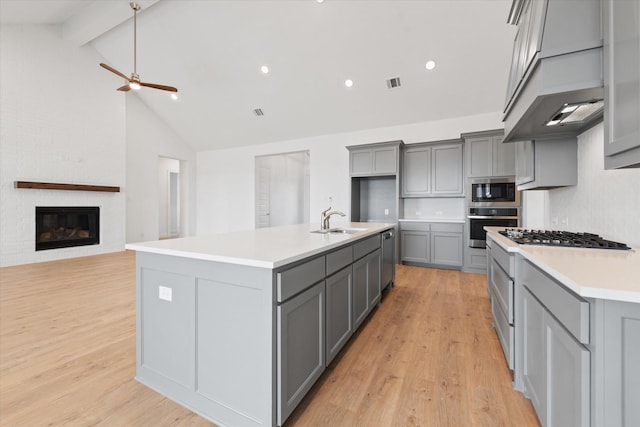 kitchen with a large fireplace, a kitchen island with sink, backsplash, gray cabinetry, and appliances with stainless steel finishes