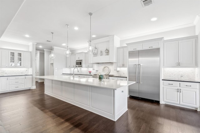 kitchen featuring built in appliances, white cabinets, a large island with sink, backsplash, and hanging light fixtures