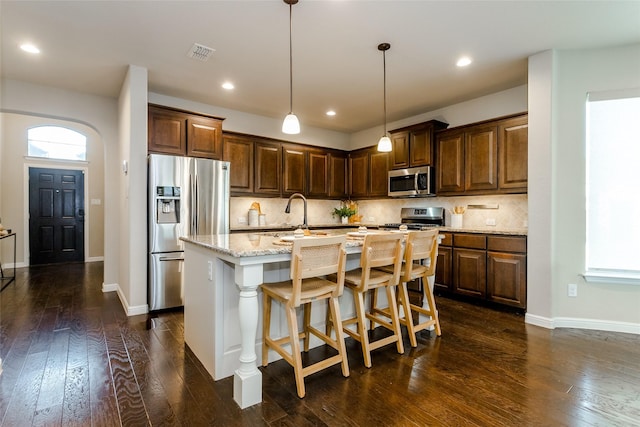 kitchen featuring a center island with sink, stainless steel appliances, light stone counters, dark hardwood / wood-style floors, and decorative light fixtures