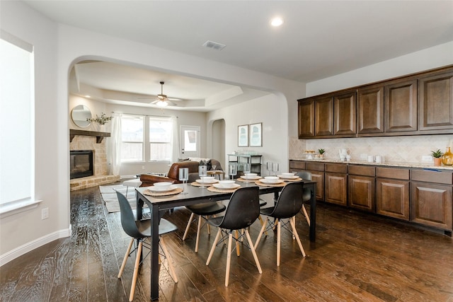 dining room with ceiling fan, a stone fireplace, a raised ceiling, and dark hardwood / wood-style floors