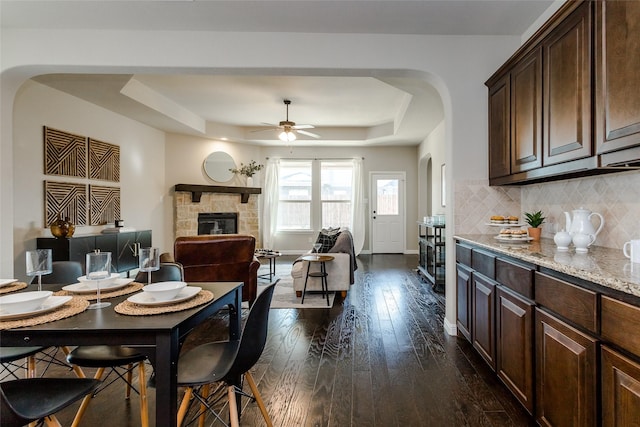 dining area with a fireplace, ceiling fan, a tray ceiling, and dark wood-type flooring