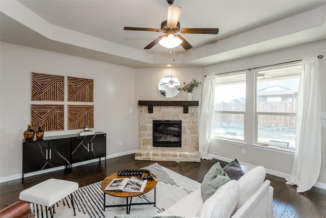 living room with dark wood-type flooring, ceiling fan, a tray ceiling, and a stone fireplace