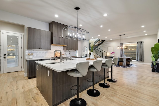 kitchen with dark brown cabinets, hanging light fixtures, an island with sink, light hardwood / wood-style floors, and backsplash