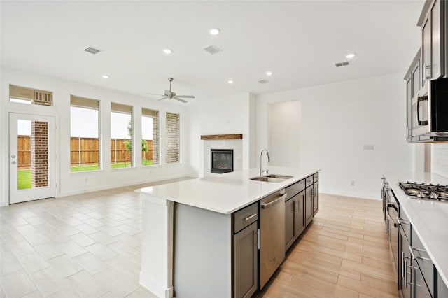 kitchen featuring appliances with stainless steel finishes, an island with sink, ceiling fan, a fireplace, and sink