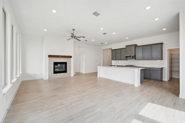 kitchen featuring a tile fireplace, ceiling fan, light hardwood / wood-style floors, an island with sink, and backsplash