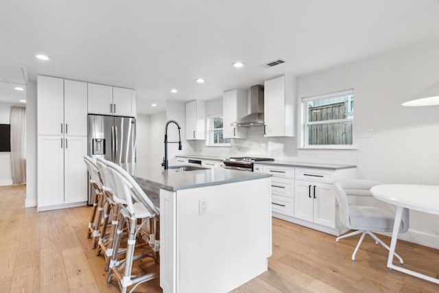 kitchen with wall chimney range hood, a kitchen island with sink, white cabinetry, appliances with stainless steel finishes, and sink