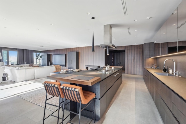 kitchen with a kitchen bar, island range hood, wooden walls, stainless steel gas cooktop, and sink