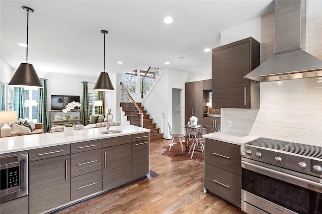 kitchen featuring hanging light fixtures, stainless steel appliances, wall chimney range hood, backsplash, and wood-type flooring