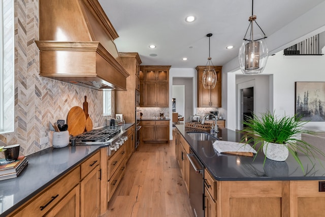 kitchen featuring a kitchen island with sink, appliances with stainless steel finishes, hanging light fixtures, custom range hood, and sink