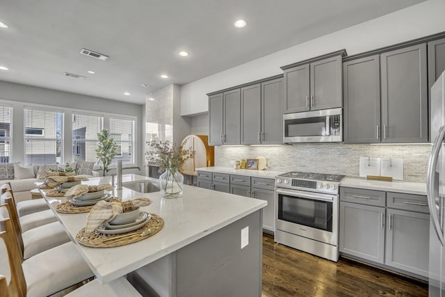 kitchen featuring a kitchen island with sink, gray cabinets, stainless steel appliances, light stone counters, and decorative backsplash