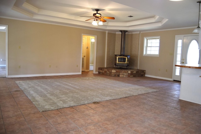 unfurnished living room featuring ceiling fan, a tray ceiling, crown molding, and a wood stove