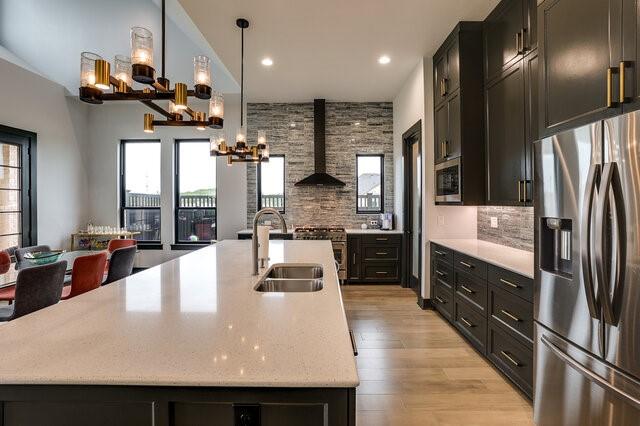 kitchen featuring wall chimney exhaust hood, an island with sink, stainless steel refrigerator with ice dispenser, decorative backsplash, and sink