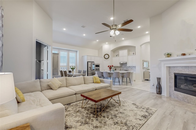 living room featuring a towering ceiling, light wood-type flooring, a tiled fireplace, and ceiling fan