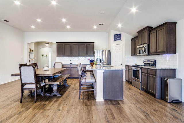 kitchen with a center island with sink, appliances with stainless steel finishes, light stone countertops, light hardwood / wood-style flooring, and backsplash