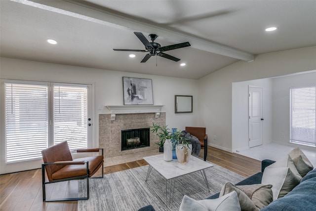 living room with ceiling fan, a tiled fireplace, light wood-type flooring, and vaulted ceiling with beams