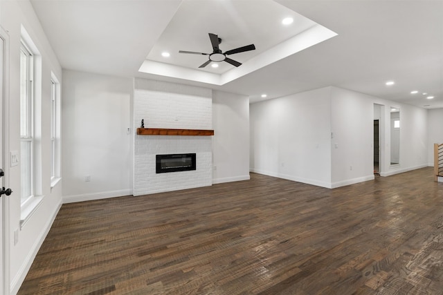 unfurnished living room featuring a brick fireplace, ceiling fan, a tray ceiling, and dark hardwood / wood-style floors