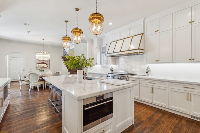 kitchen with a kitchen island with sink, appliances with stainless steel finishes, custom range hood, pendant lighting, and white cabinets