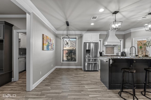 kitchen featuring stainless steel fridge with ice dispenser, custom range hood, white cabinetry, stone counters, and a kitchen breakfast bar