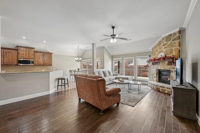 living room featuring a stone fireplace, vaulted ceiling, ornamental molding, dark wood-type flooring, and ceiling fan with notable chandelier