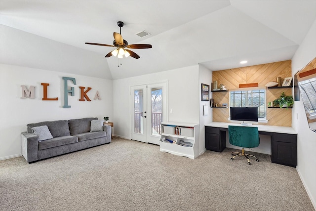 living room featuring built in desk, wooden walls, vaulted ceiling, ceiling fan, and light colored carpet