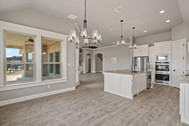 kitchen with light stone counters, a center island with sink, appliances with stainless steel finishes, ceiling fan with notable chandelier, and white cabinets