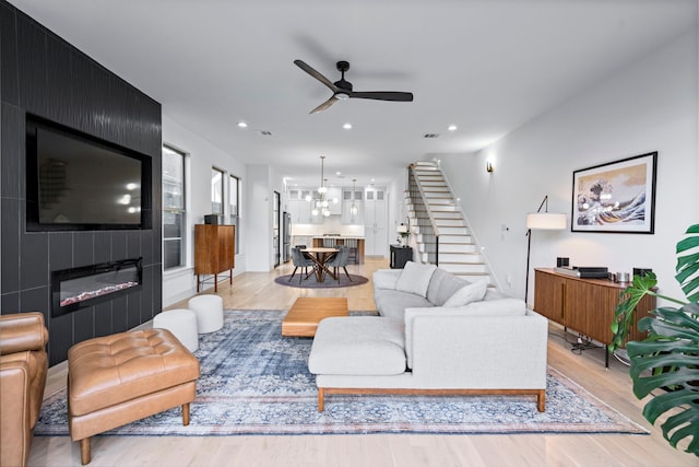 living room with a large fireplace, light wood-type flooring, and ceiling fan with notable chandelier