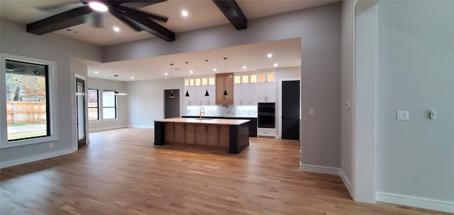 kitchen featuring hanging light fixtures, light wood-type flooring, a kitchen island with sink, white cabinetry, and beamed ceiling