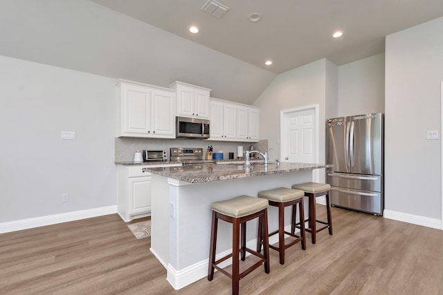 kitchen featuring stainless steel appliances, an island with sink, light wood-type flooring, light stone counters, and white cabinetry