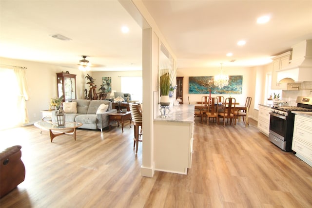 kitchen featuring white cabinets, light hardwood / wood-style flooring, ceiling fan with notable chandelier, stainless steel gas range oven, and premium range hood