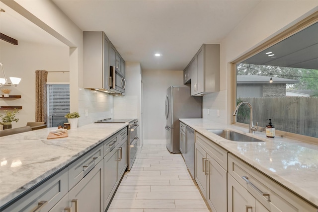 kitchen with gray cabinets, stainless steel appliances, light stone countertops, sink, and backsplash