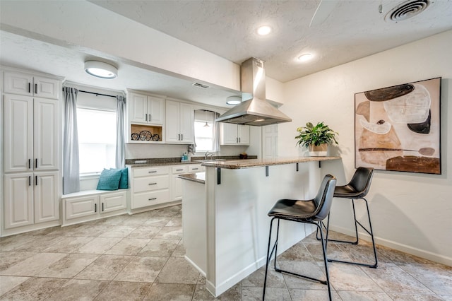 kitchen featuring kitchen peninsula, a textured ceiling, island exhaust hood, white cabinetry, and a kitchen breakfast bar