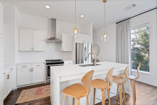 kitchen with wall chimney exhaust hood, decorative light fixtures, stainless steel appliances, an island with sink, and white cabinetry