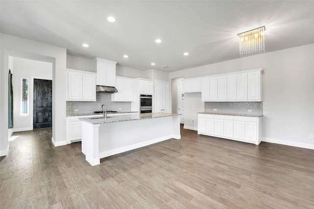 kitchen with hardwood / wood-style floors, a kitchen island with sink, light stone countertops, white cabinets, and tasteful backsplash