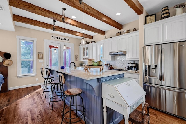 kitchen with hanging light fixtures, stainless steel refrigerator with ice dispenser, a kitchen island with sink, white cabinets, and beamed ceiling