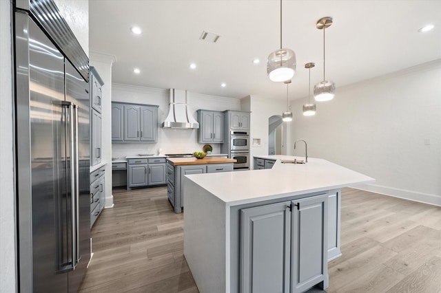 kitchen with appliances with stainless steel finishes, sink, wall chimney range hood, an island with sink, and wood counters