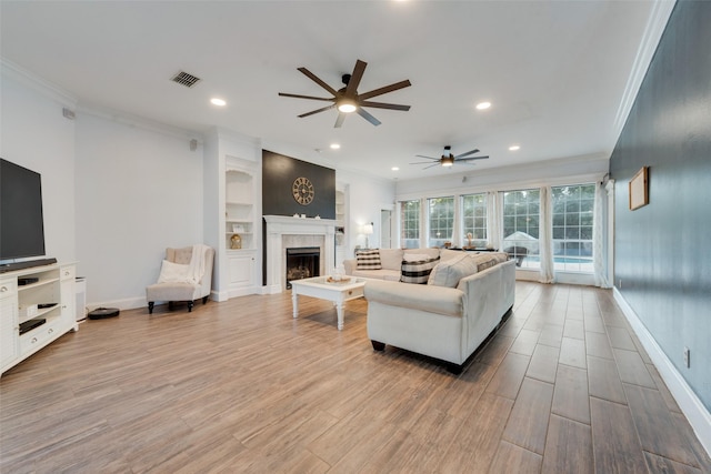 living room with built in features, a tiled fireplace, ceiling fan, and crown molding