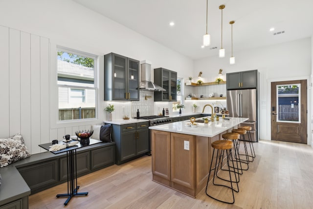 kitchen featuring light hardwood / wood-style floors, appliances with stainless steel finishes, a kitchen island with sink, hanging light fixtures, and wall chimney exhaust hood