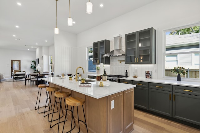 kitchen with wall chimney exhaust hood, light hardwood / wood-style flooring, sink, hanging light fixtures, and a center island with sink