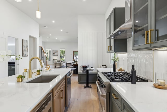 kitchen with stainless steel appliances, decorative light fixtures, wall chimney range hood, light stone counters, and sink