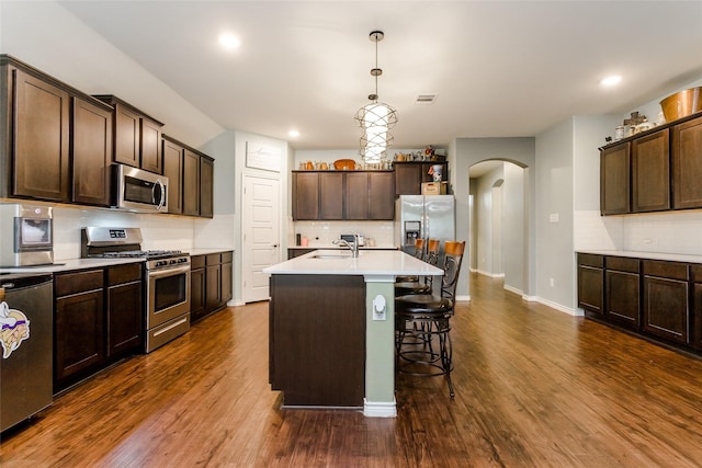 kitchen featuring dark hardwood / wood-style flooring, stainless steel appliances, hanging light fixtures, a center island with sink, and dark brown cabinets